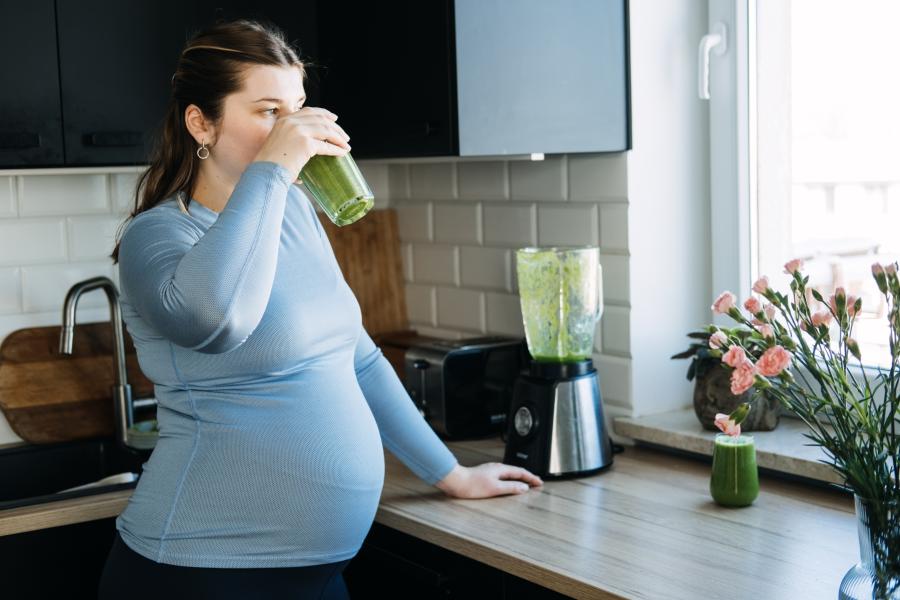A pregnant woman stands in the kitchen drinking a green smoothie.